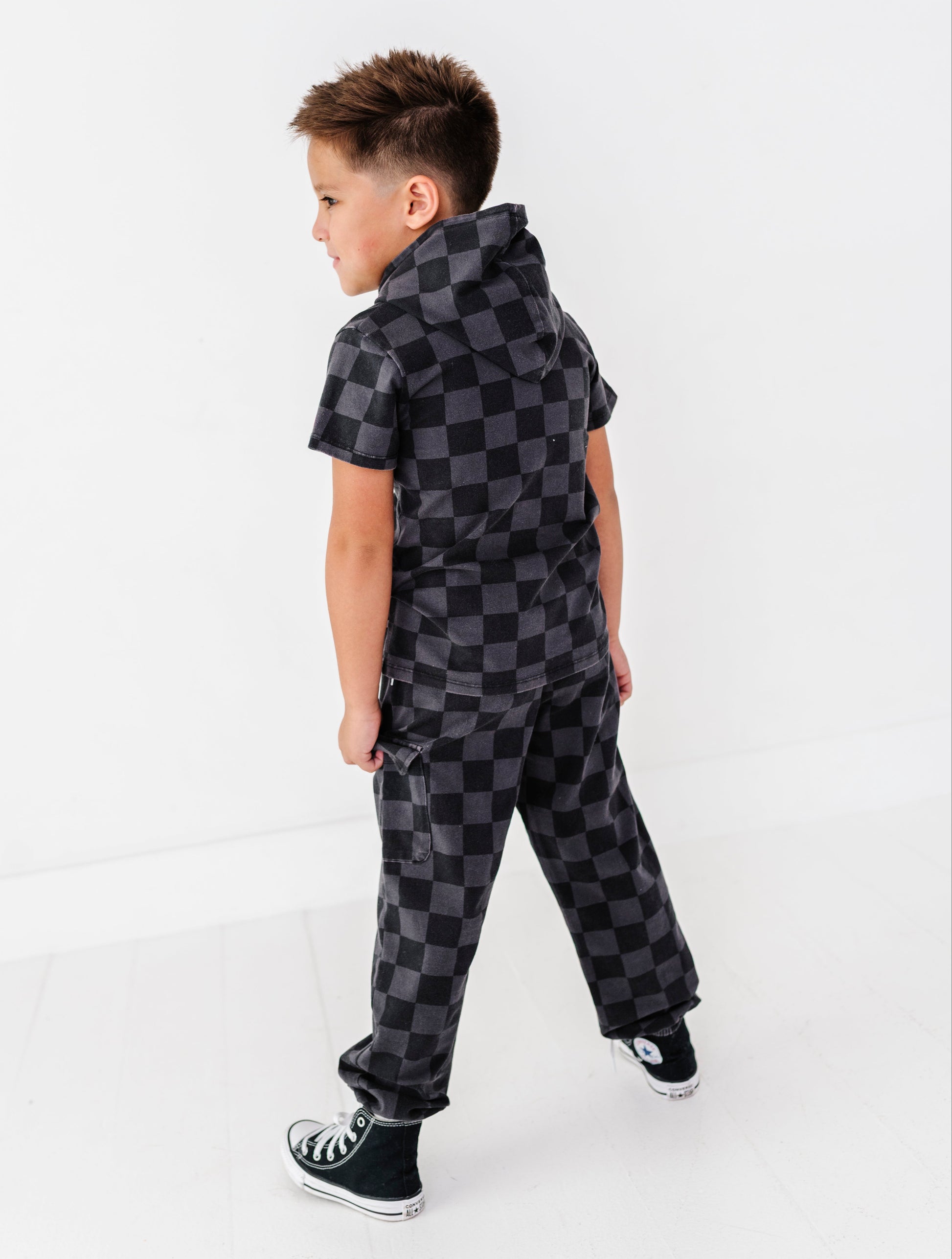 A young boy wearing a black and gray checkered short-sleeve hooded jumpsuit, shown from a side-back angle against a clean white studio background.