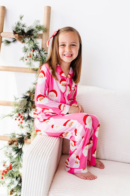 A young girl with a pink Santa hat sits on a white ribbed sofa wearing a pink and white Santa Claus patterned bamboo pajama set. To her left is a wooden ladder decorated with green pine garlands and red berries.