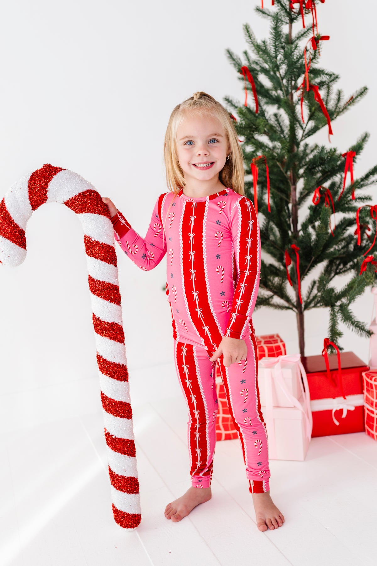 A young girl stands barefoot in a white room wearing a pink and red holiday candy cane print bamboo two-piece pajama set, holding a giant red and white candy cane prop next to a decorated Christmas tree and wrapped gifts.