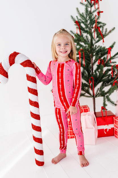 A young girl stands barefoot in a white room wearing a pink and red holiday candy cane print bamboo two-piece pajama set, holding a giant red and white candy cane prop next to a decorated Christmas tree and wrapped gifts.