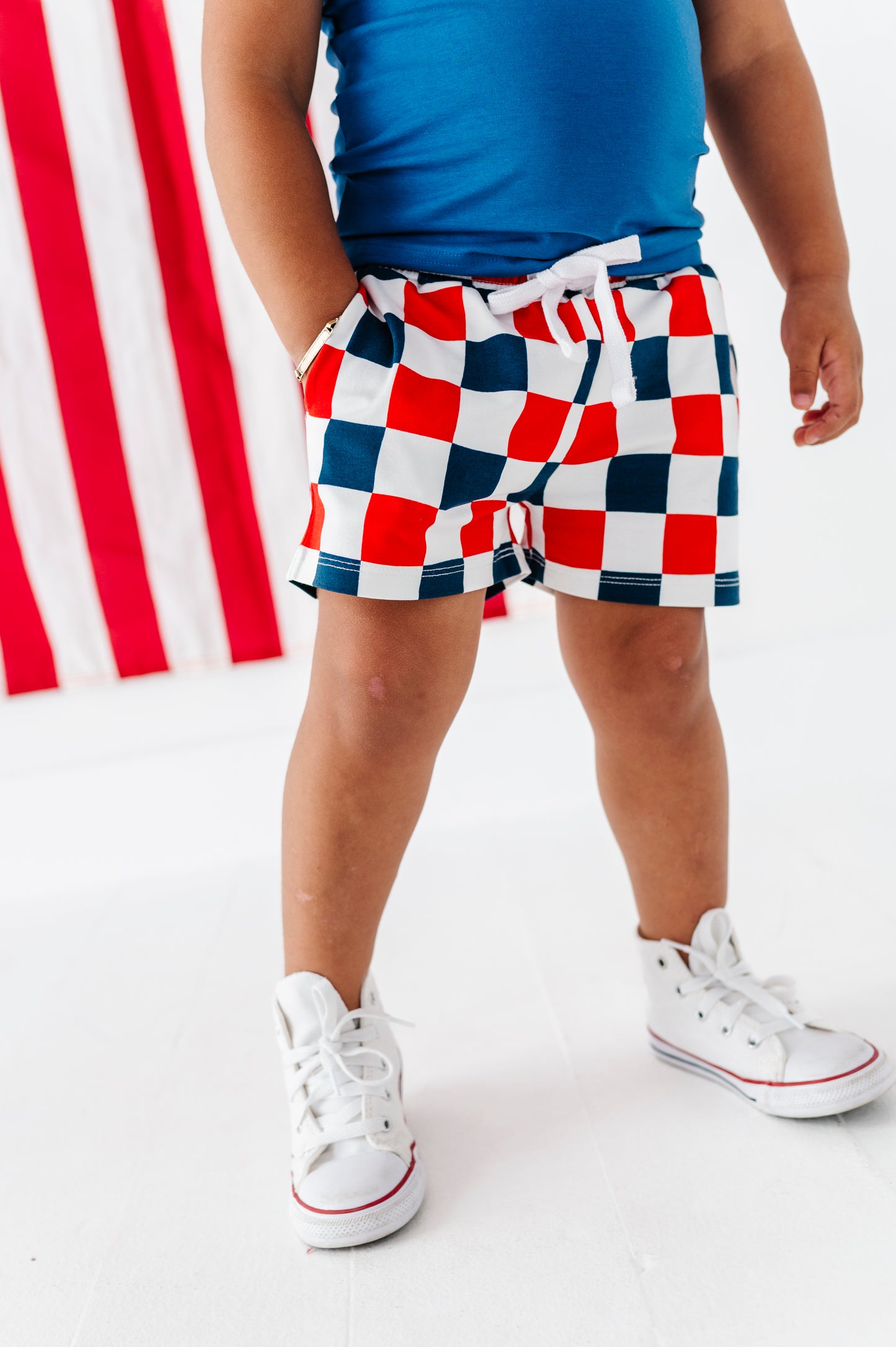 Young boy wearing red, white, and blue checkered bamboo shorts with a blue top and white sneakers on a white background.