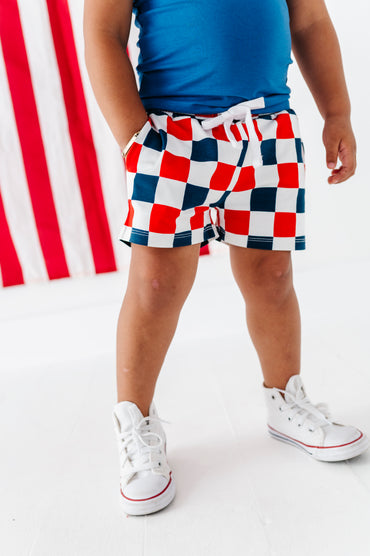 Young boy wearing red, white, and blue checkered bamboo shorts with a blue top and white sneakers on a white background.