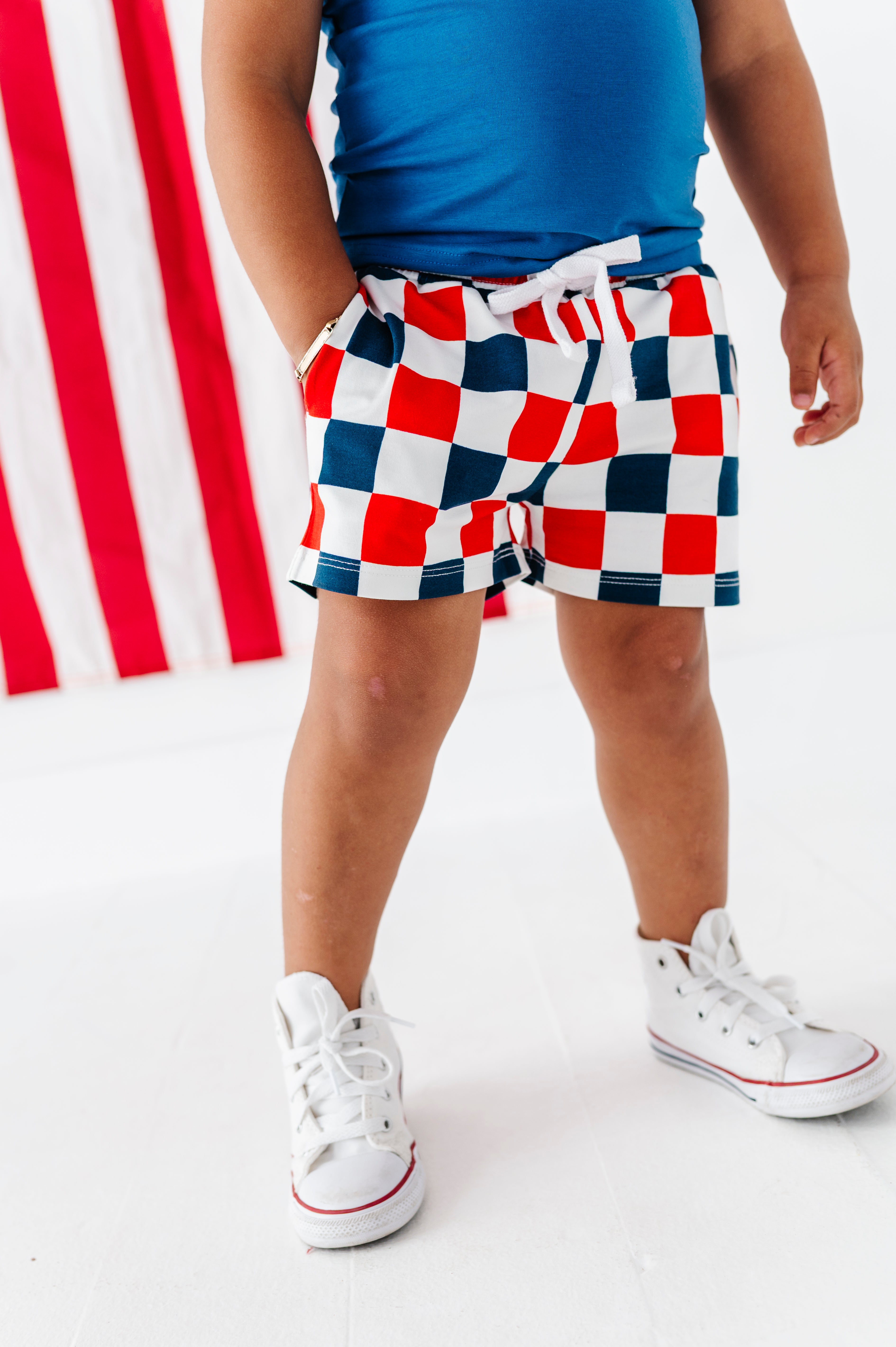 Young boy wearing red, white, and blue checkered bamboo shorts with a blue top and white sneakers on a white background.