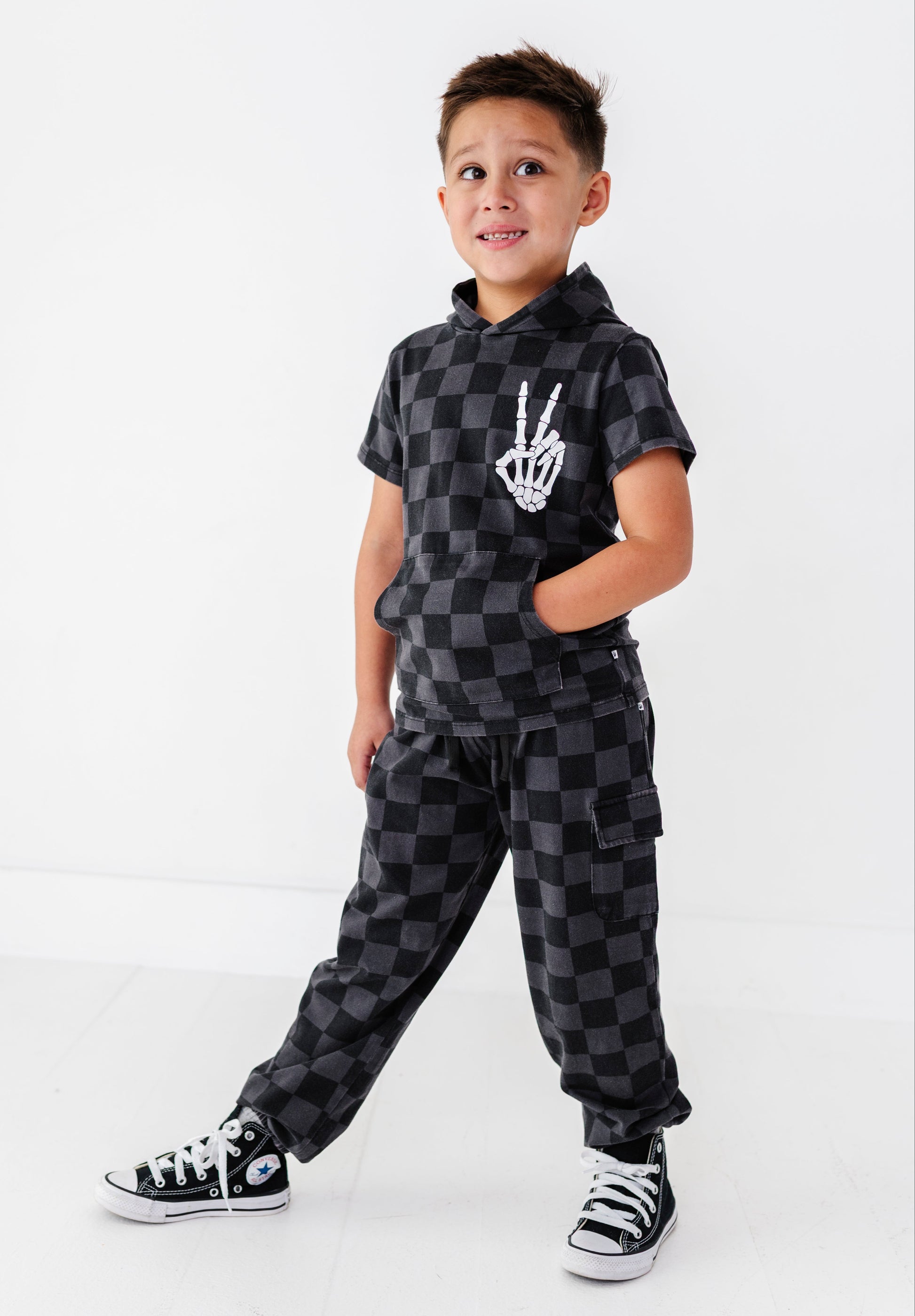 A young boy modeling a black and gray checkered two-piece outfit consisting of a short-sleeve hoodie with a white skeleton hand graphic and matching cargo pants, paired with black and white sneakers, against a plain white studio background.