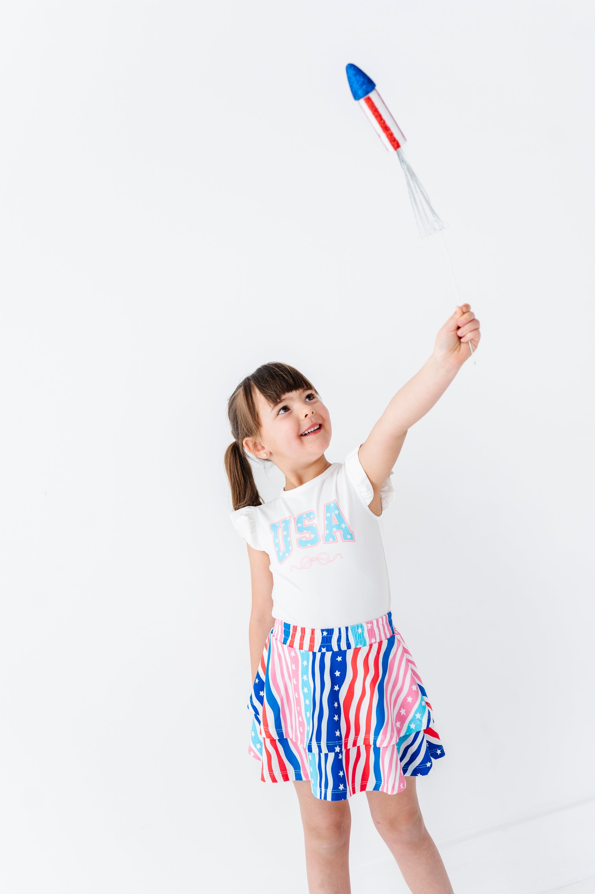 Child wearing a 'USA' bamboo shirt and American flag skort, holding a toy.