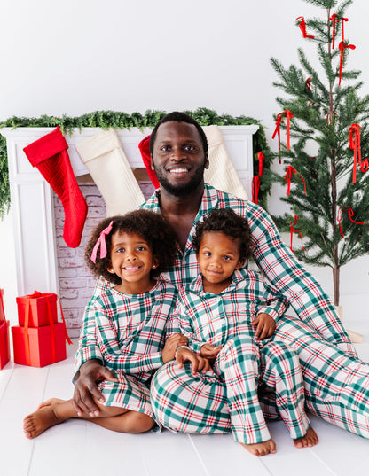A dad and his two little ones, all wearing matching classic Christmas plaid pajamas and sitting in front of a Christmas tree