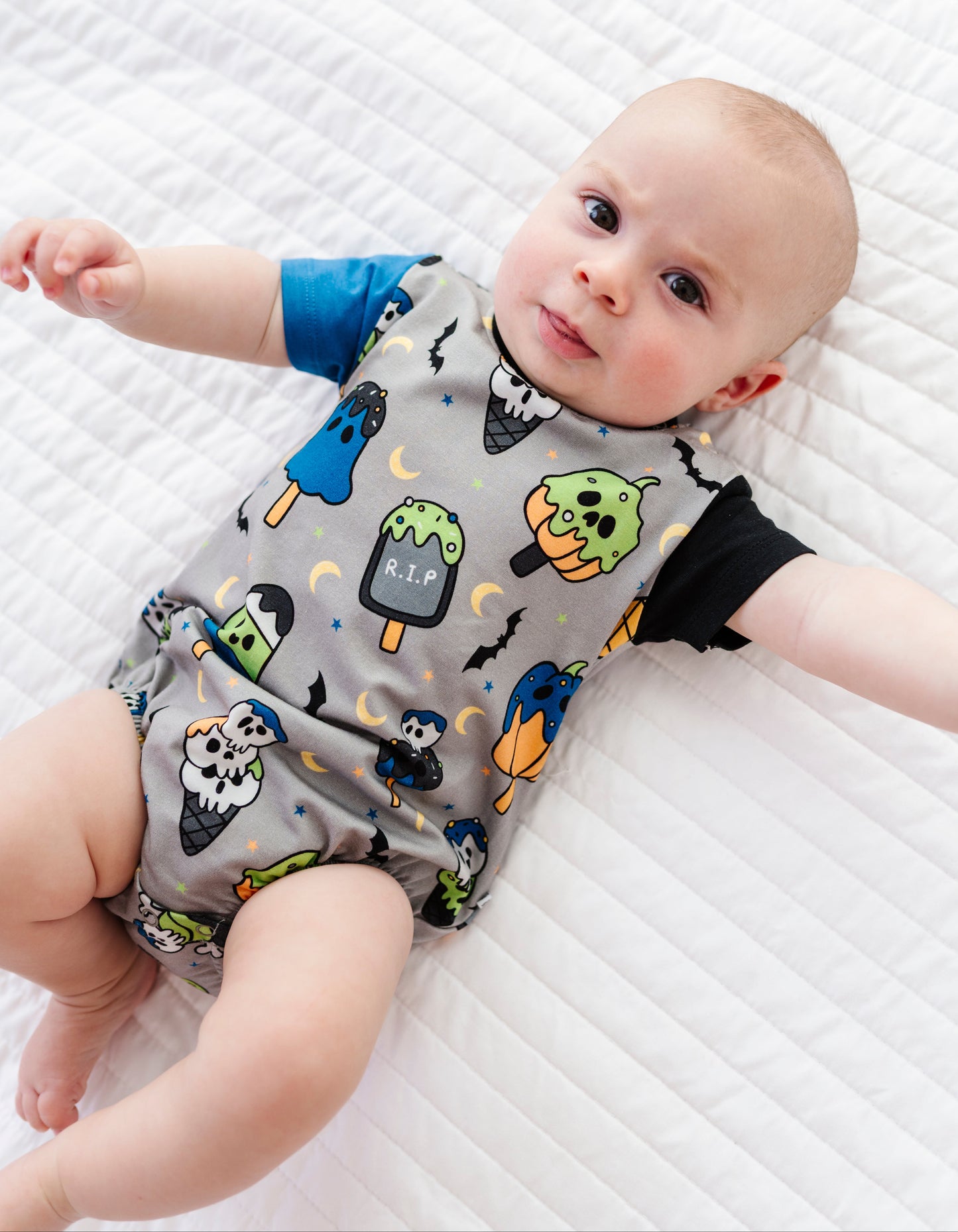 A baby lying on a white quilted mattress wearing a gray short-sleeve romper with a colorful Halloween-themed print of ghosts, pumpkins, bats, and moons, with blue and black sleeves.