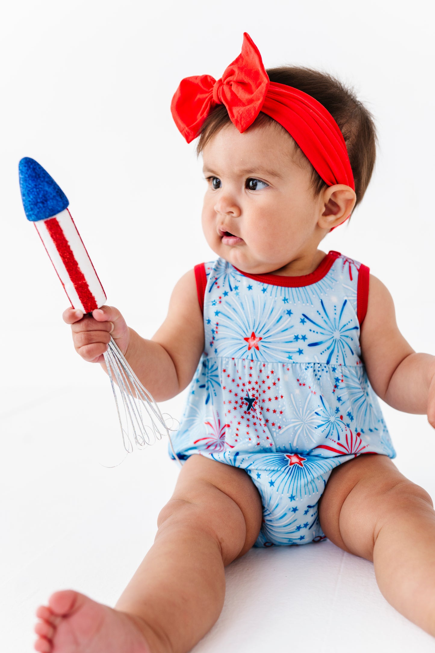 Baby wearing a red, white, and blue bamboo bubble romper with a firework pattern, holding a toy rocket.