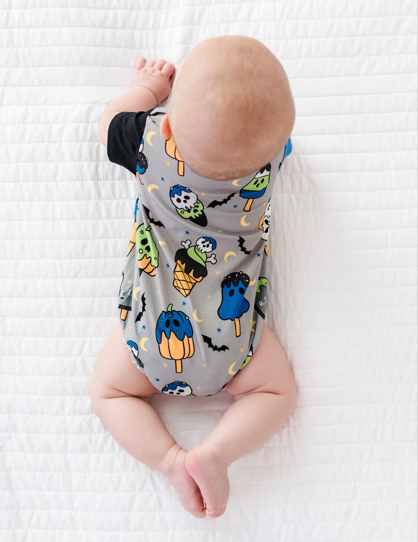 A baby lying on their stomach on a white textured blanket, wearing a gray short-sleeve bodysuit printed with colorful popsicle illustrations.