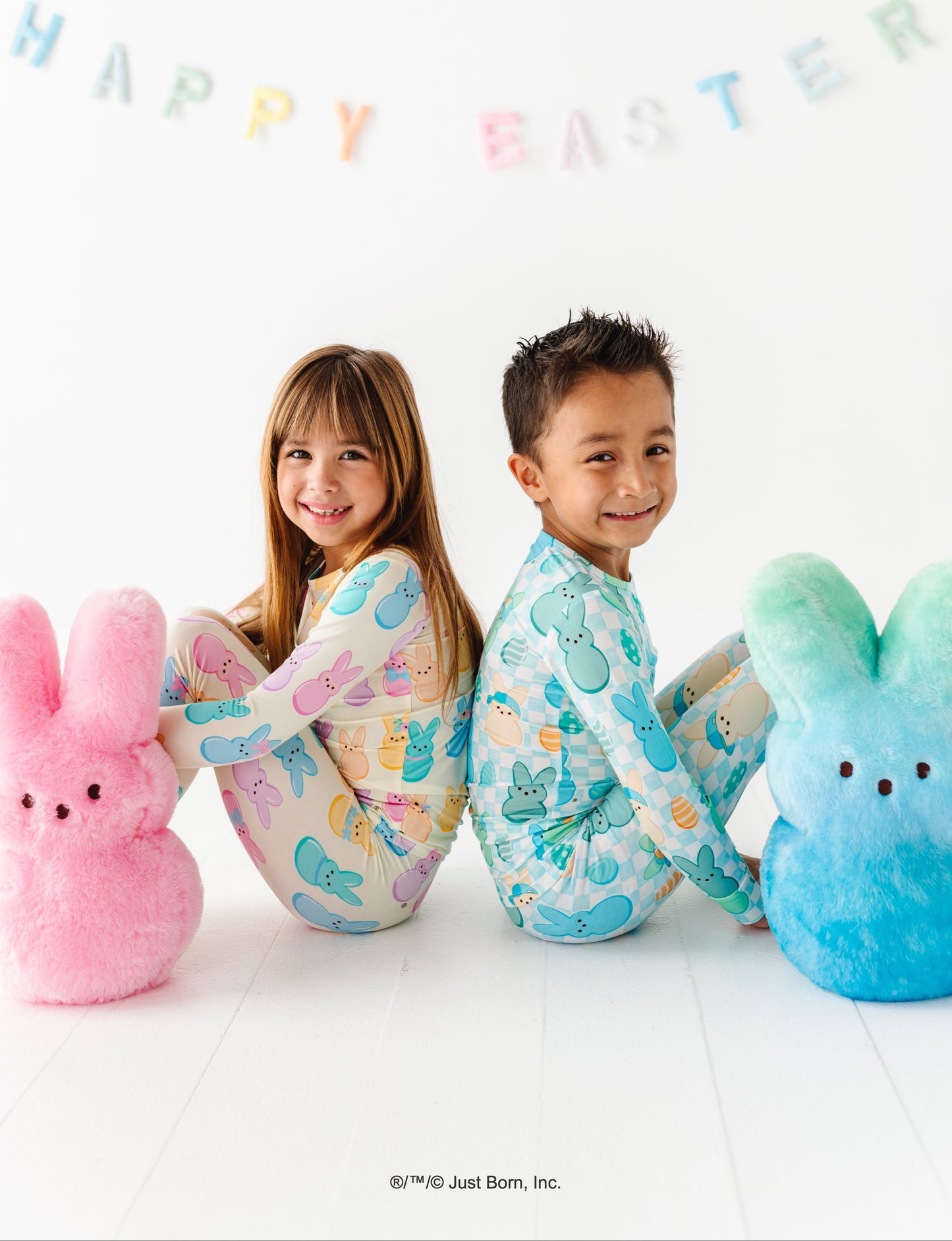 Two children wearing pajamas with bunny ears, sitting between two large Peeps bunny plush, with 'Happy Easter' text in the background.