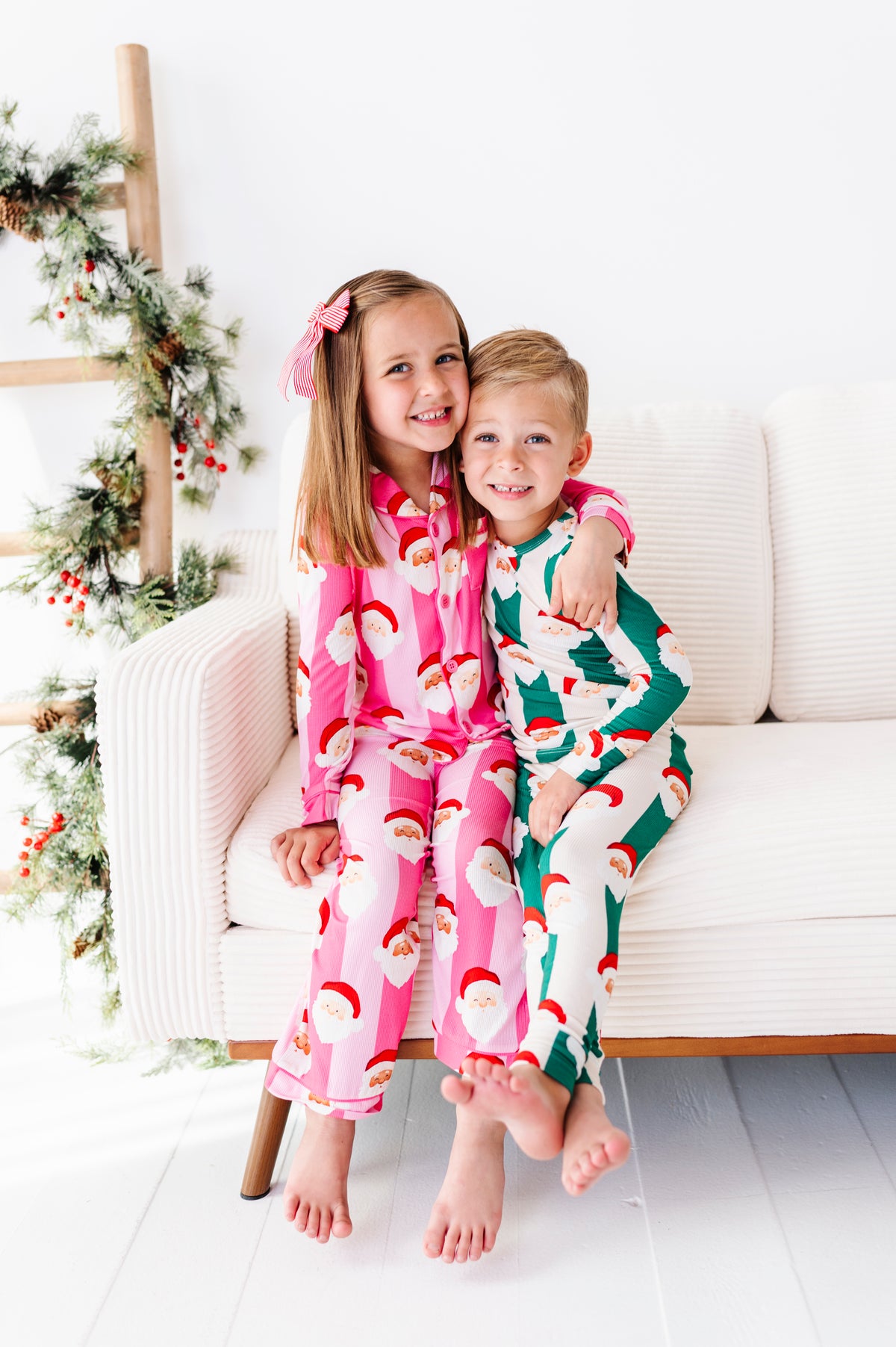 Two children sitting on a white sofa wearing matching Christmas bamboo pajamas with Santa Claus patterns, one in pink and one in green, in a bright holiday-themed room.