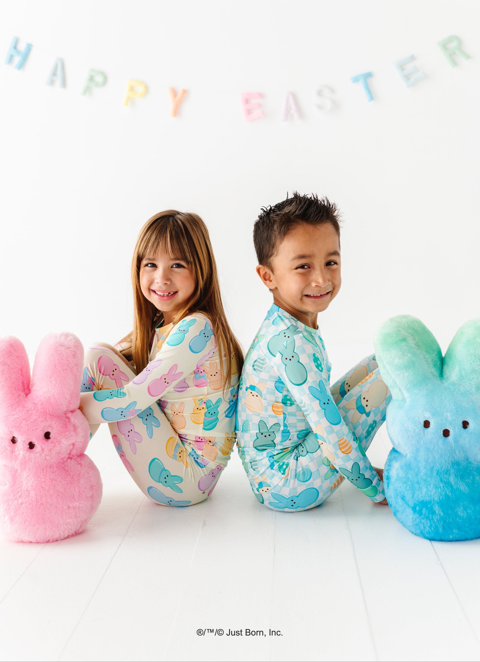 Two children wearing pajamas with sitting between two large Peeps bunny plush toys, with 'Happy Easter' text in the background.