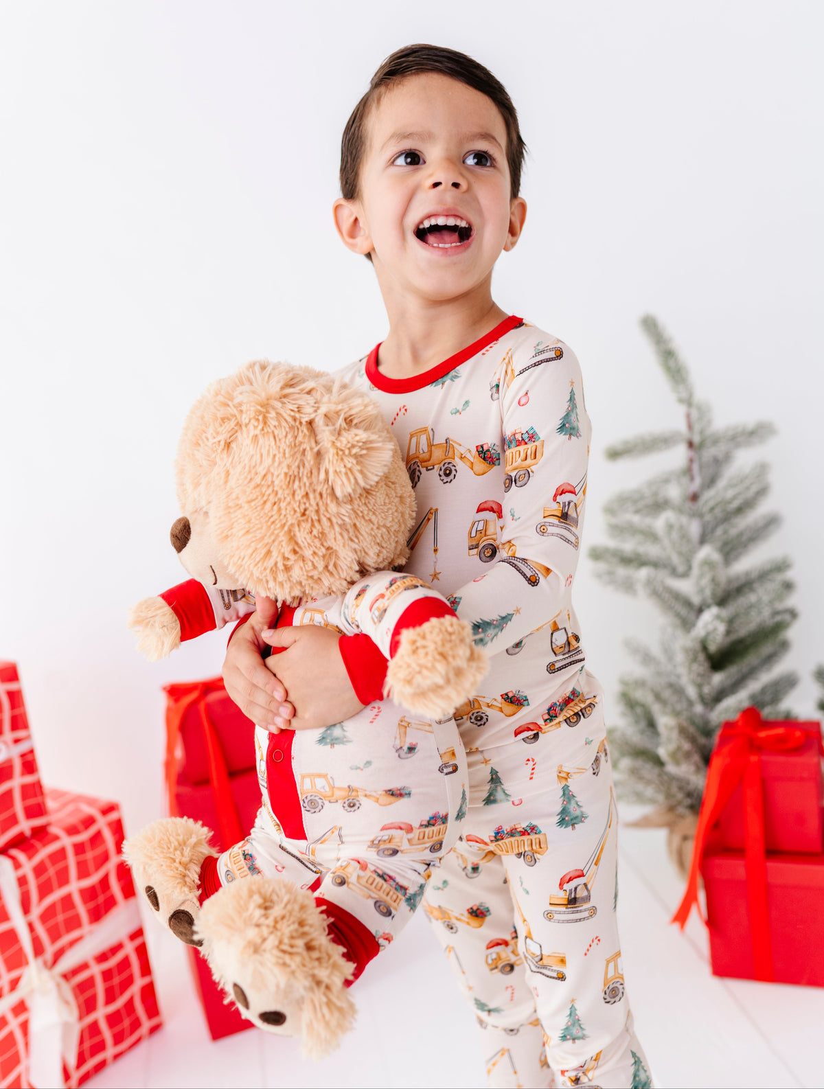 A young boy wearing Christmas construction-themed two-piece pajamas, holding a teddy bear wearing a matching construction bamboo romper 