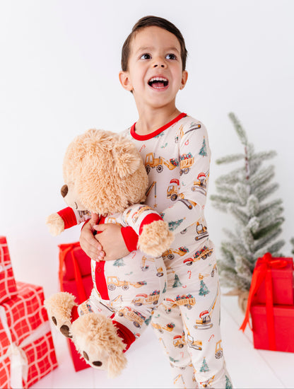 A young boy wearing Christmas construction-themed two-piece pajamas, holding a teddy bear wearing a matching construction bamboo romper 