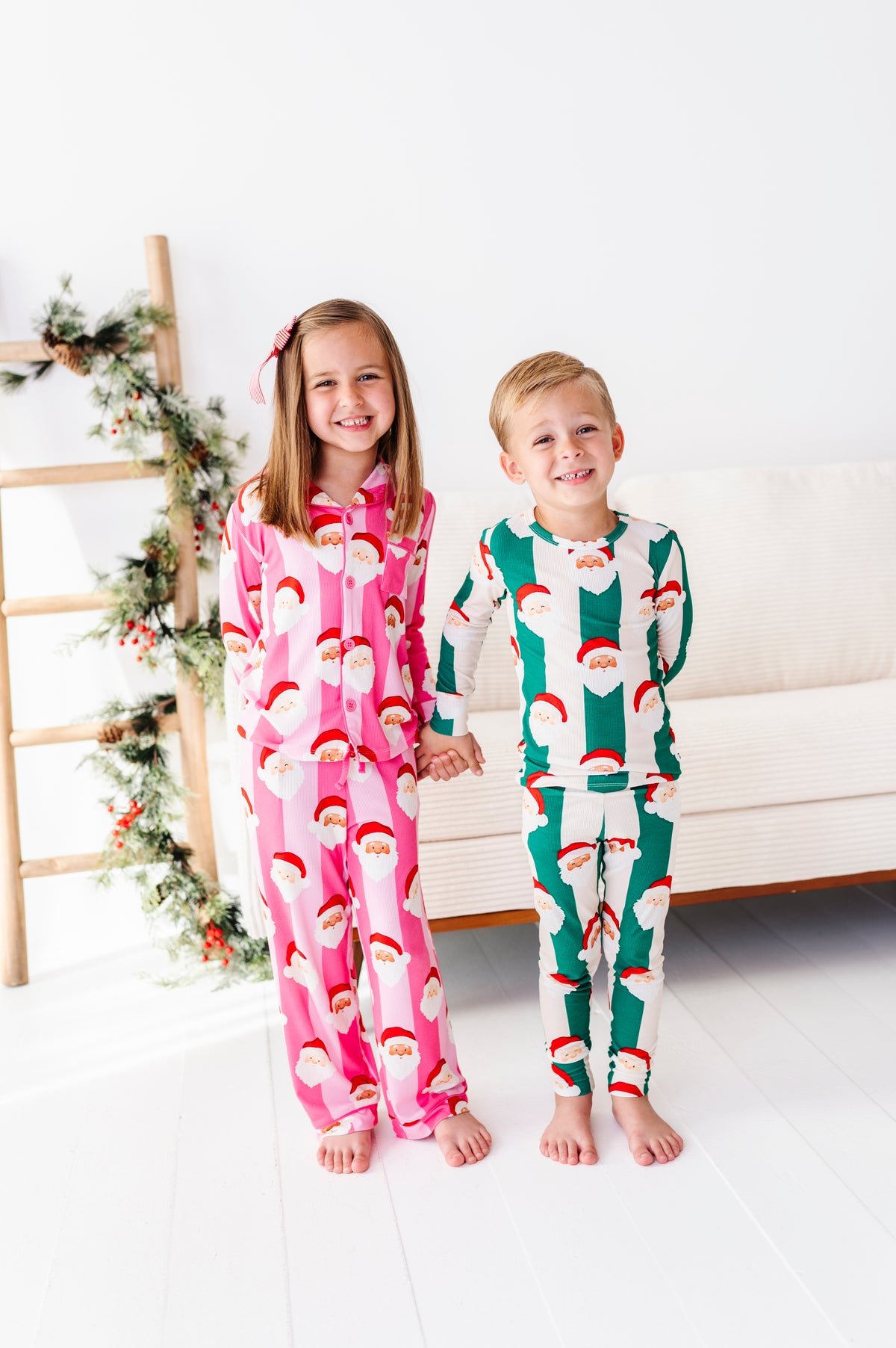 A young girl and boy standing barefoot indoors, wearing matching Christmas-themed long-sleeve bamboo pajamas. The girl's pajamas are pink with a white Santa Claus face print, and the boy's pajamas are green with a white Santa Claus face print.
