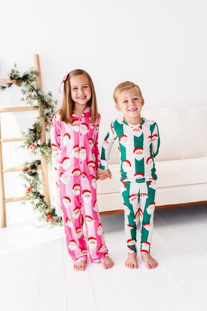 A young girl and boy standing barefoot indoors, wearing matching Christmas-themed long-sleeve bamboo pajamas. The girl's pajamas are pink with a white Santa Claus face print, and the boy's pajamas are green with a white Santa Claus face print.