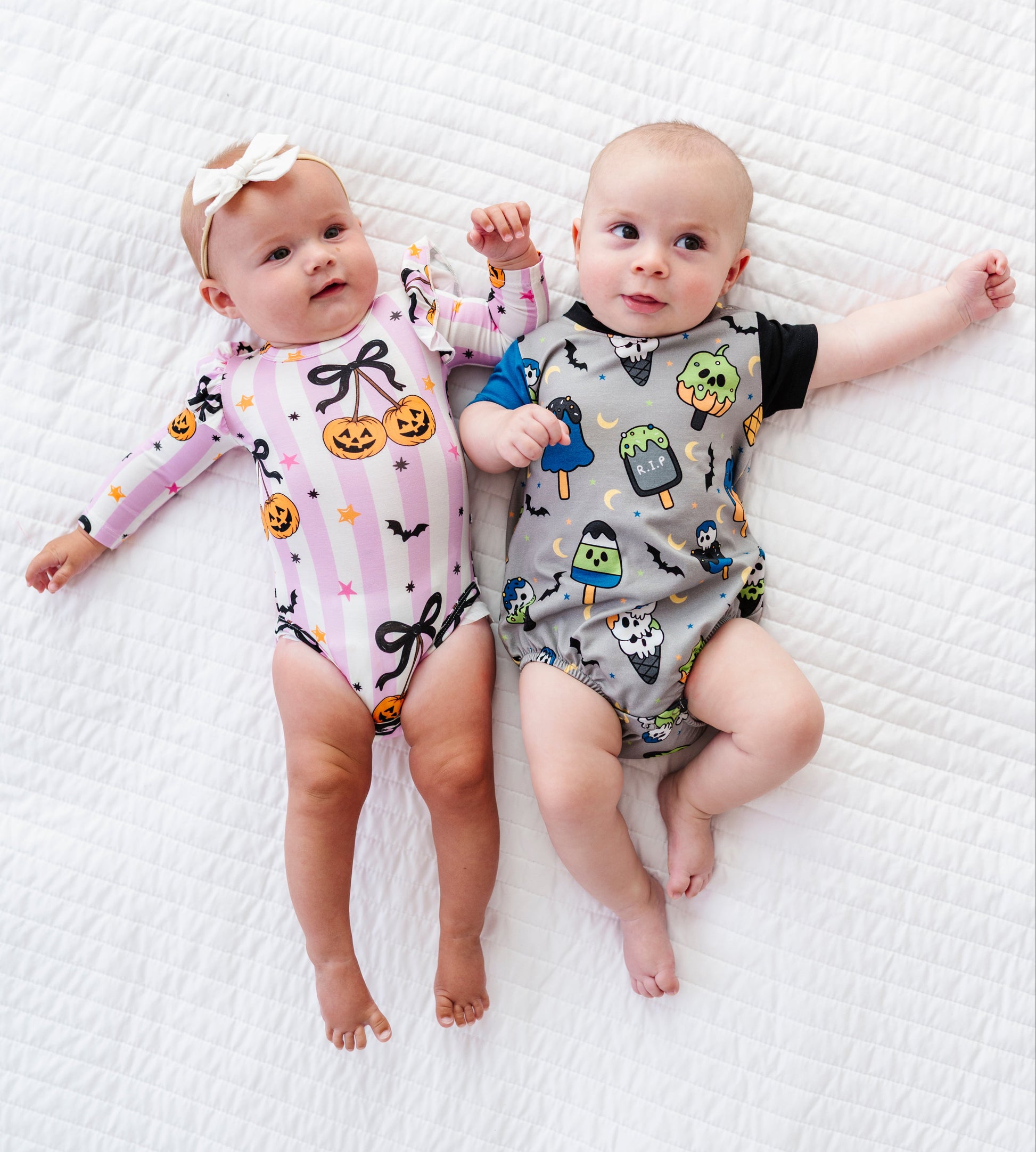 Two babies lying side by side on a white quilted blanket, wearing matching Halloween-themed bodysuits. The baby on the left wears a light pink long-sleeve bodysuit with black bows and orange jack-o'-lantern prints, paired with a white bow headband.