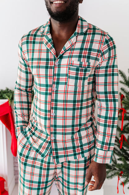 A close-up detail shot of a man wearing a bamboo lounge set in a classic Christmas plaid print, standing in front of a fireplace and a Christmas tree