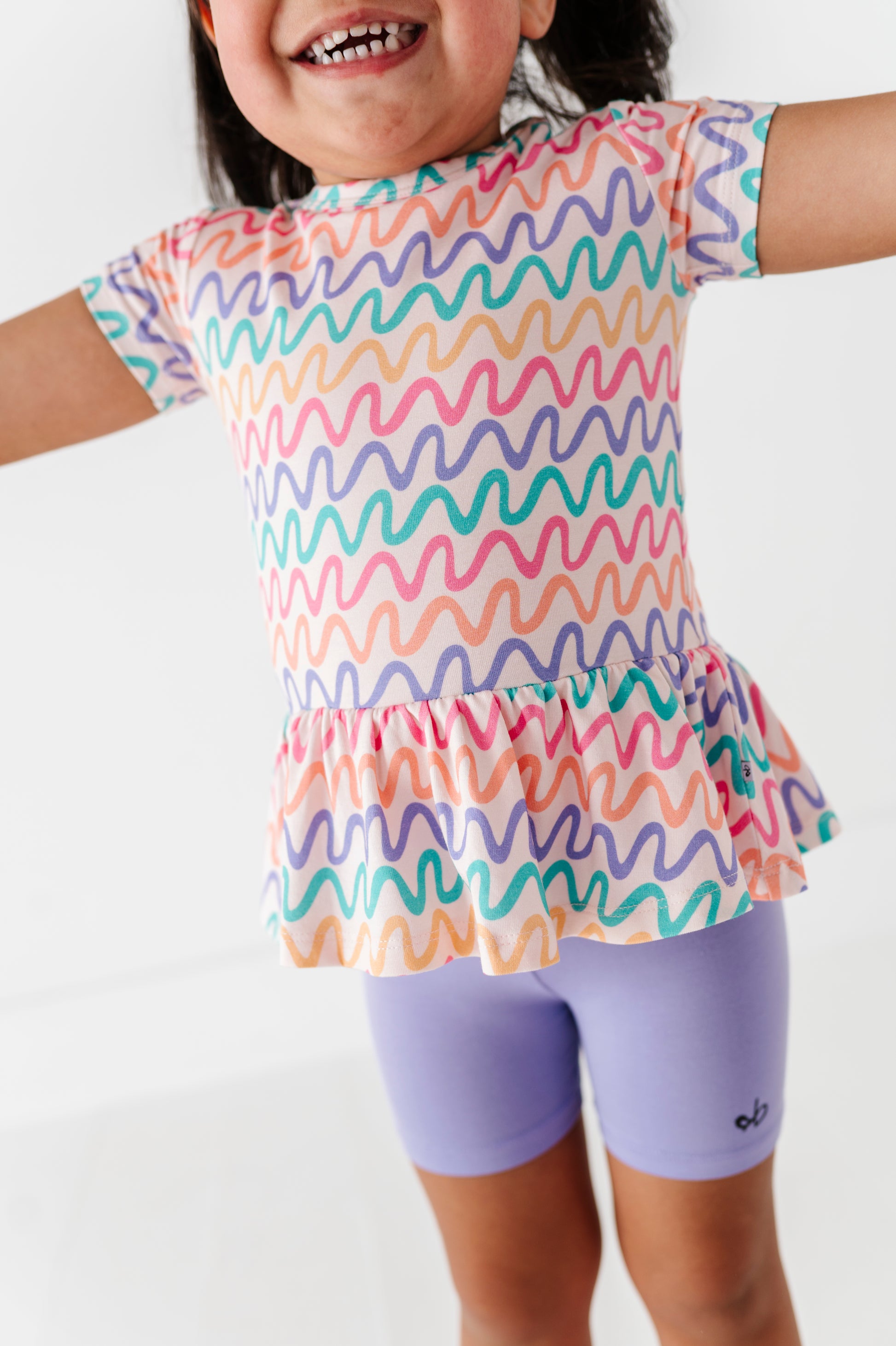 Child wearing a colorful wavy patterned bamboo/cotton top and purple bamboo biker shorts on a white background