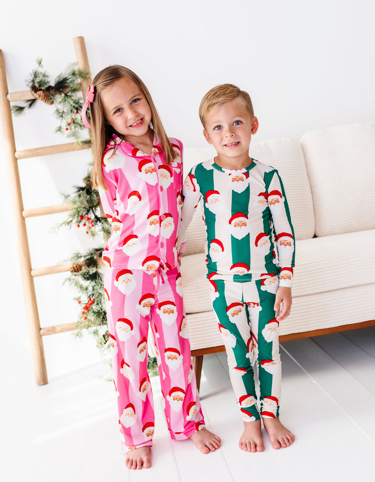 A brother and sister holding hands while wearing coordinating Christmas bamboo pajamas featuring green stripes, pink stripes, and cute Santa faces 