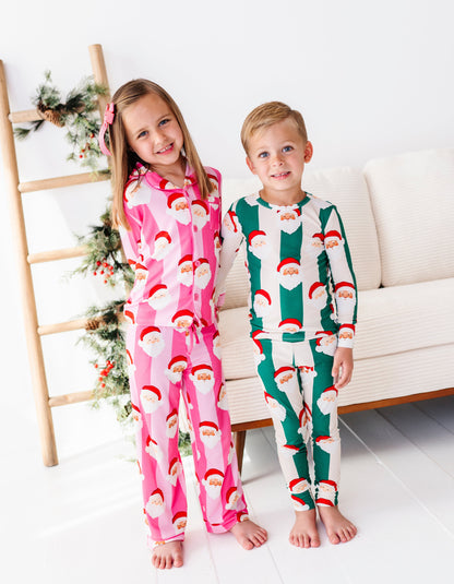 A brother and sister holding hands while wearing coordinating Christmas bamboo pajamas featuring green stripes, pink stripes, and cute Santa faces 