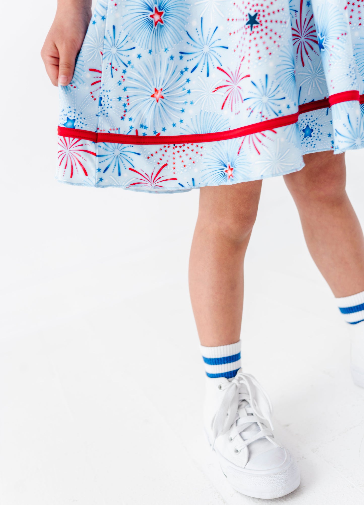 Little girl wearing a bamboo dress with fireworks pattern, white sneakers, and striped socks on a white background