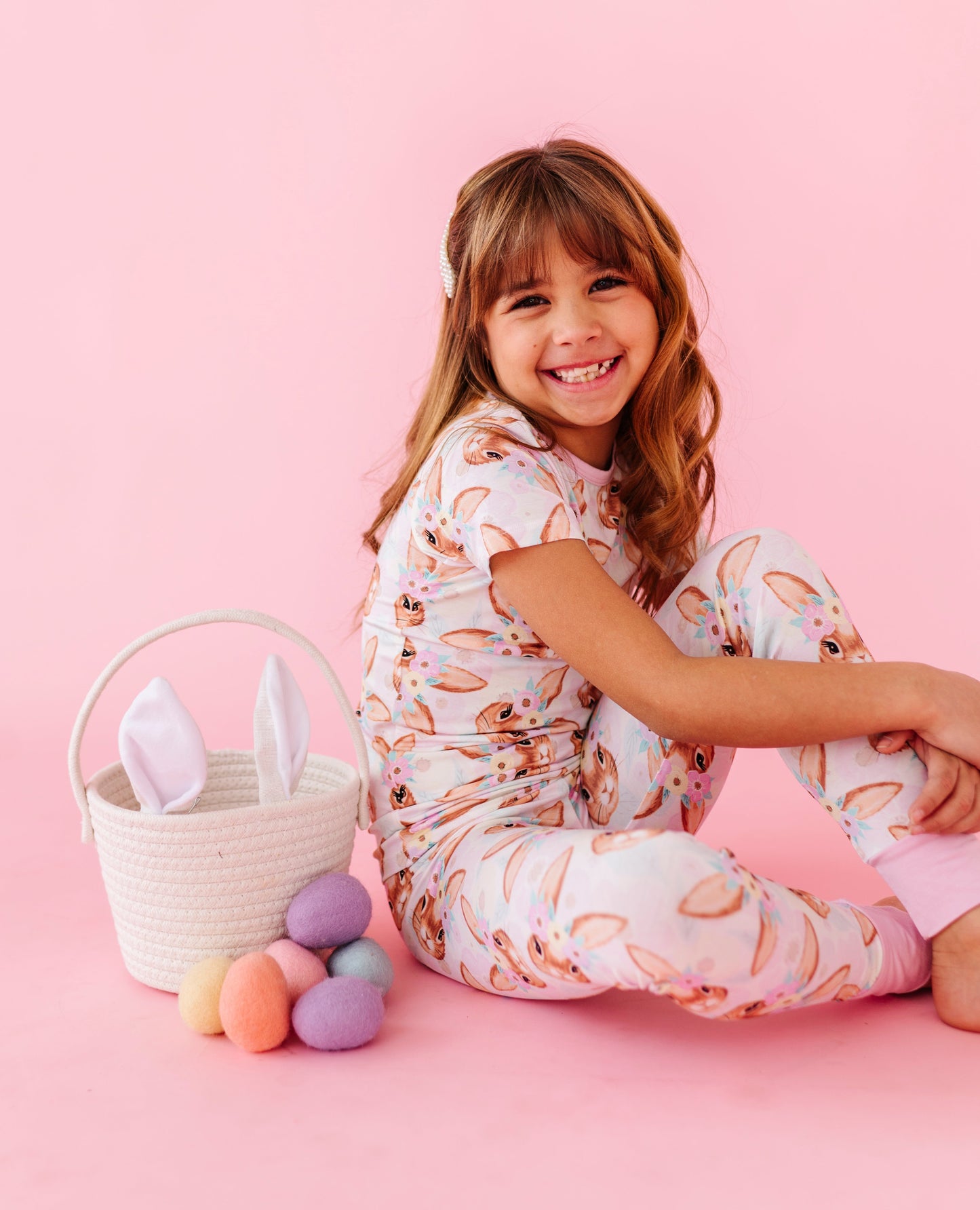 Young girl in bamboo pajamas sitting on a pink floor with Easter eggs and a basket.
