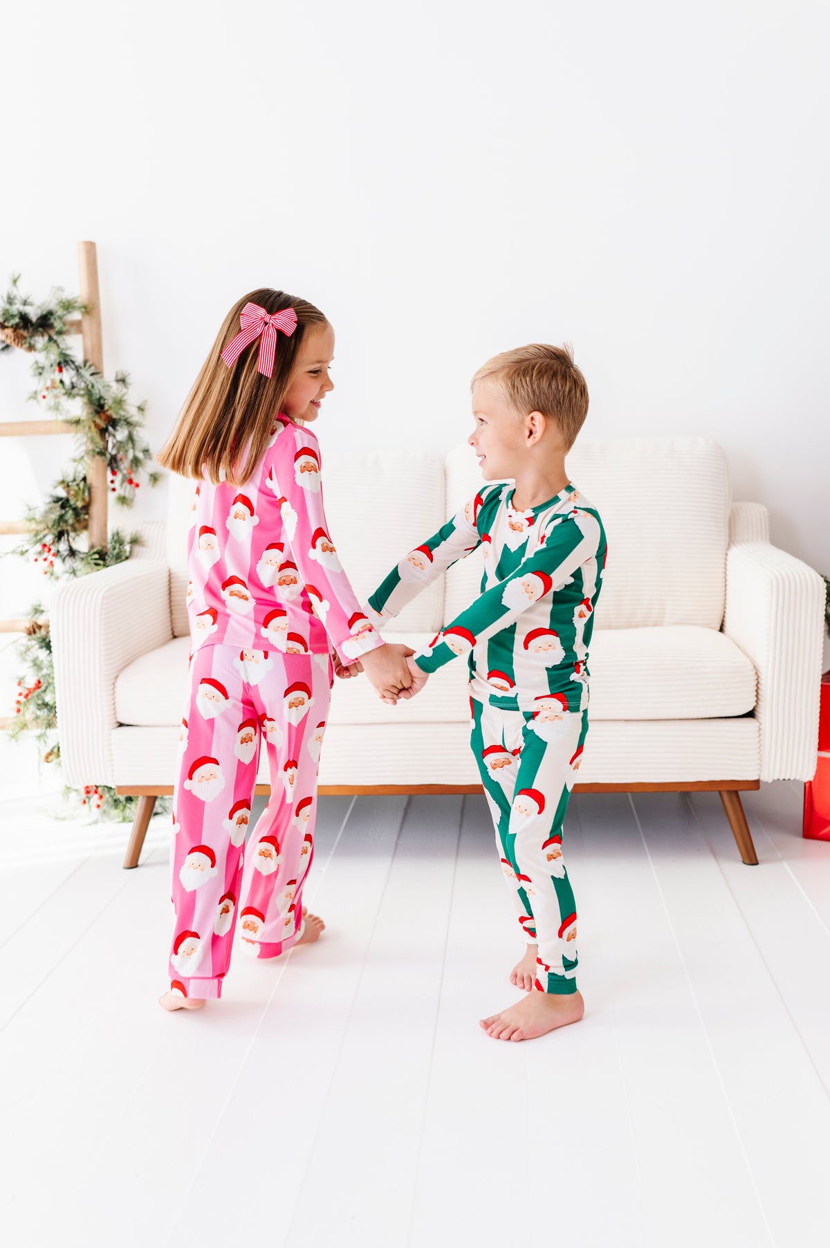 Two children standing and holding hands in a bright white room, wearing matching Christmas bamboo pajamas. The girl's pajamas are pink with a Santa Claus face print, and the boy's pajamas are green with a Santa Claus face print.