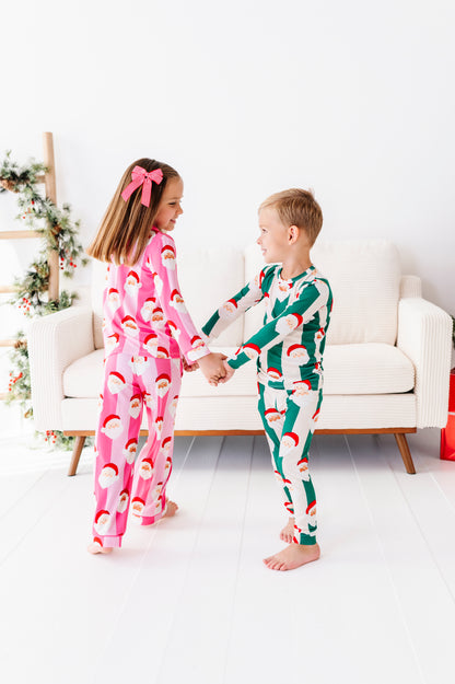 Two children standing and holding hands in a bright white room, wearing matching Christmas bamboo pajamas. The girl's pajamas are pink with a Santa Claus face print, and the boy's pajamas are green with a Santa Claus face print.
