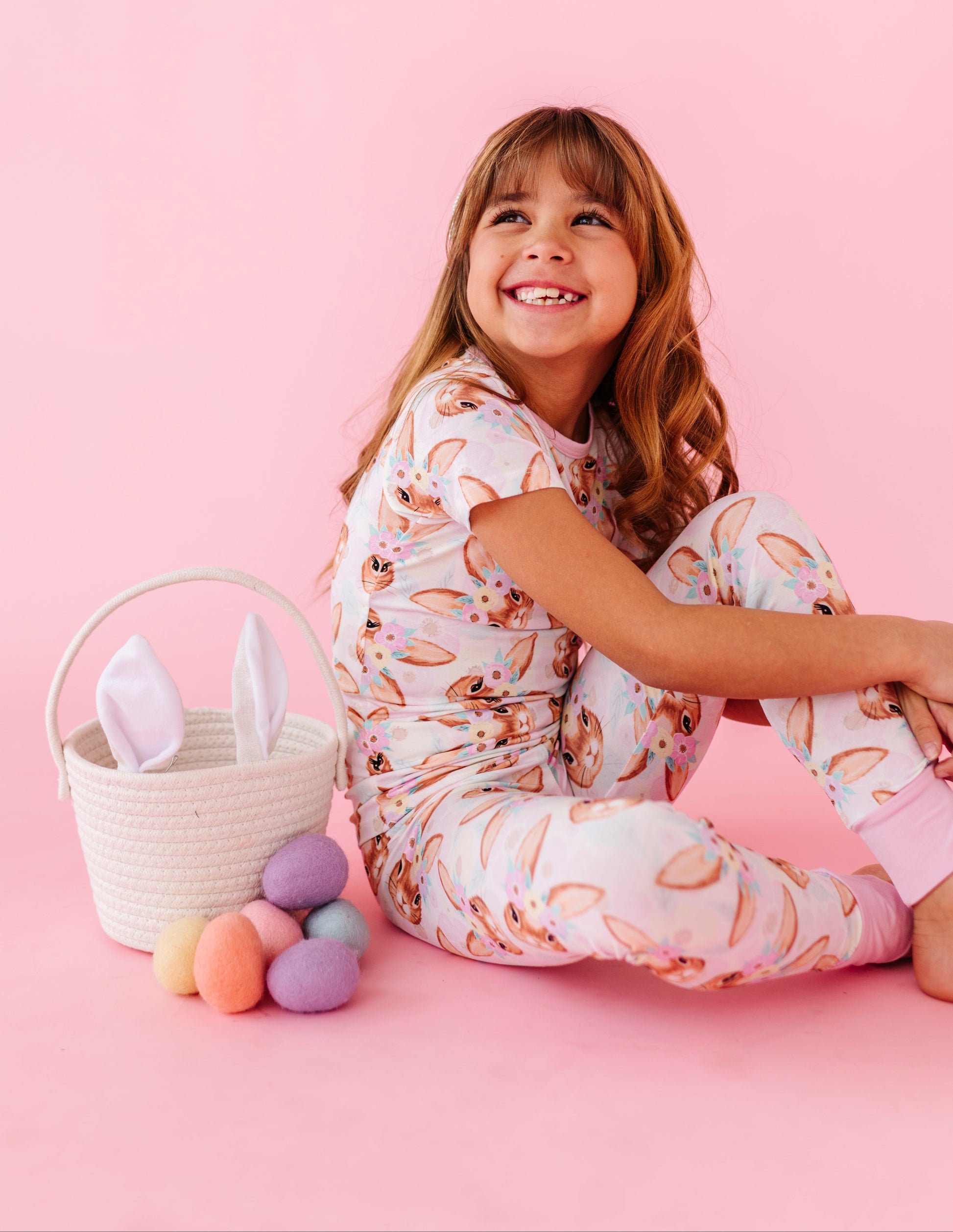 Child in a floral and bunny bamboo pajamas set sitting on a pink floor with Easter eggs and a basket.