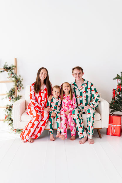 A family of four wearing matching Christmas-themed bamboo pajamas sits together on a white sofa in a bright, modern room. The mother wears a red and white Santa pattern, the father wears a green and white Santa pattern, and the two children wear matching pajamas in pink and green.