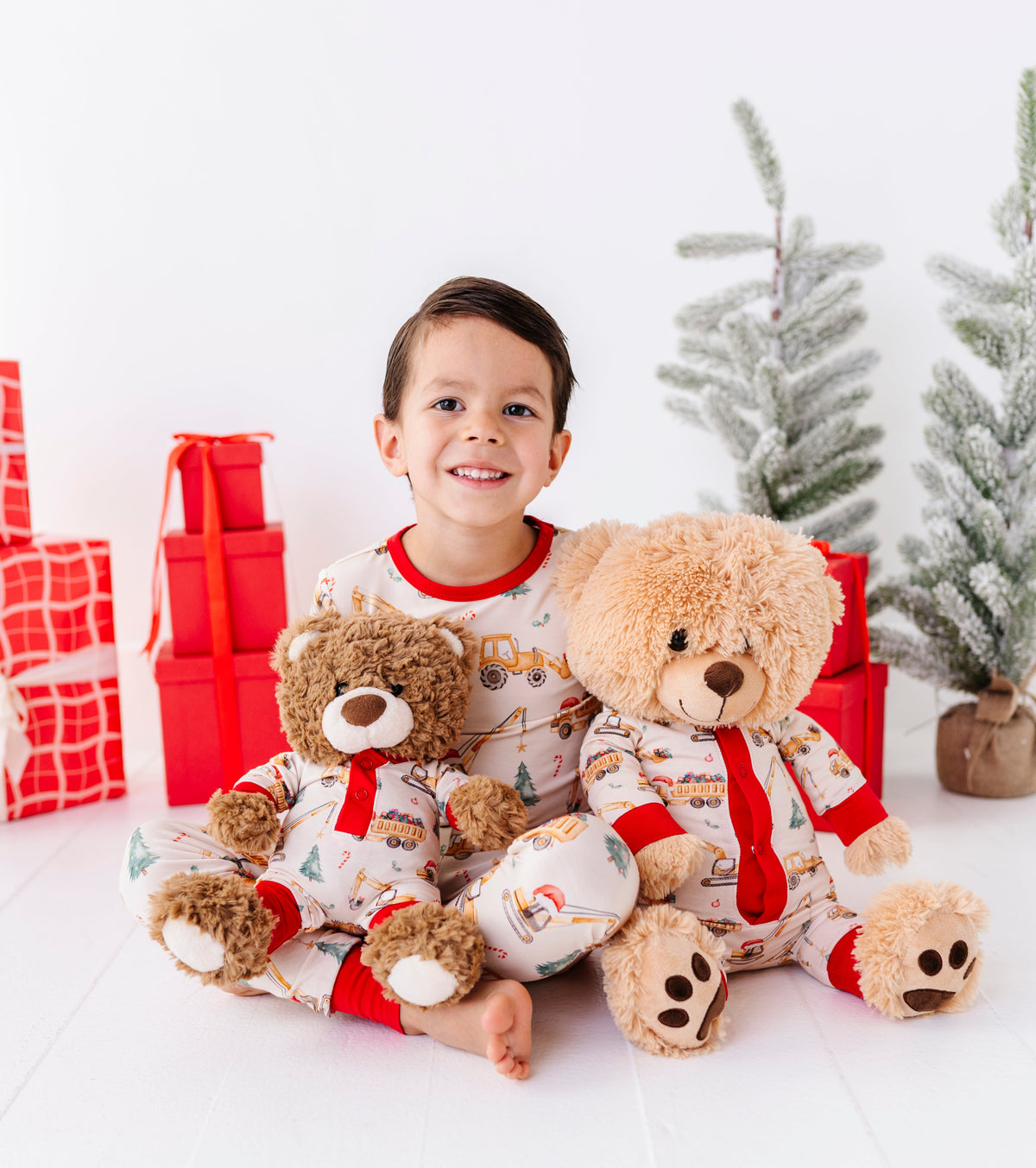 A little boy wearing a Christmas construction-themed two-piece bamboo pajama set while holding two teddy bears wearing matching construction rompers 