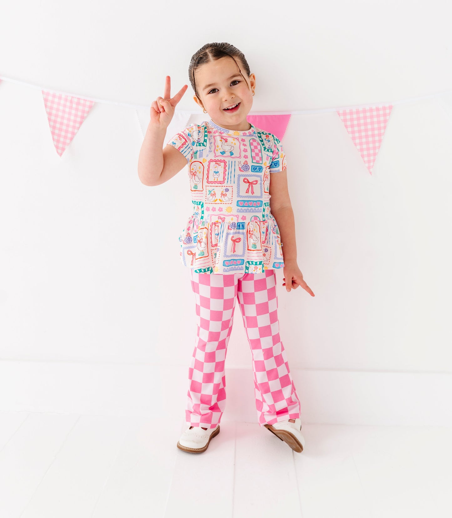 Child wearing a colorful outfit with pink checkered pants, standing against a white background with small flags.