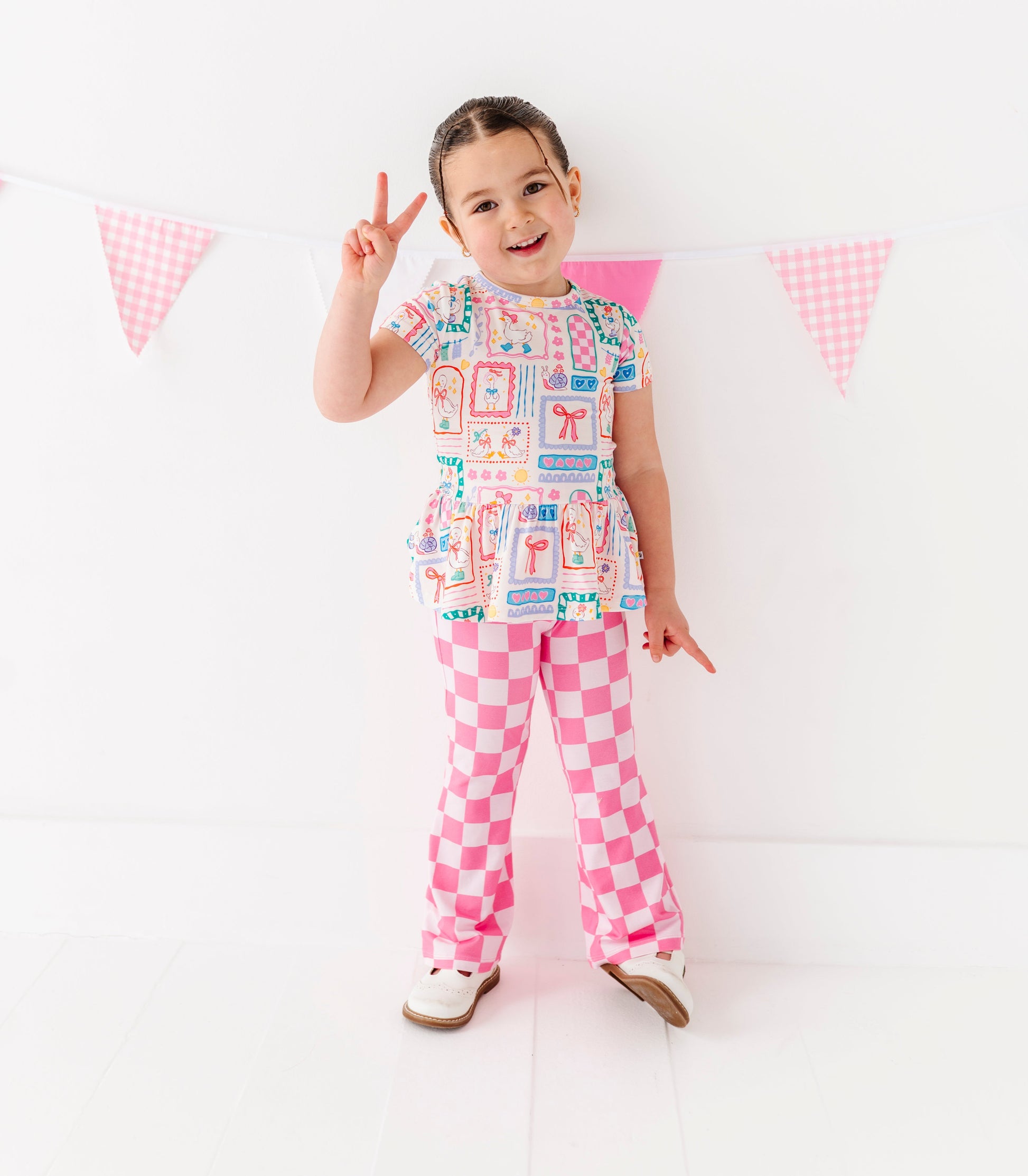 Child wearing a colorful outfit with pink checkered pants, standing against a white background with small flags.