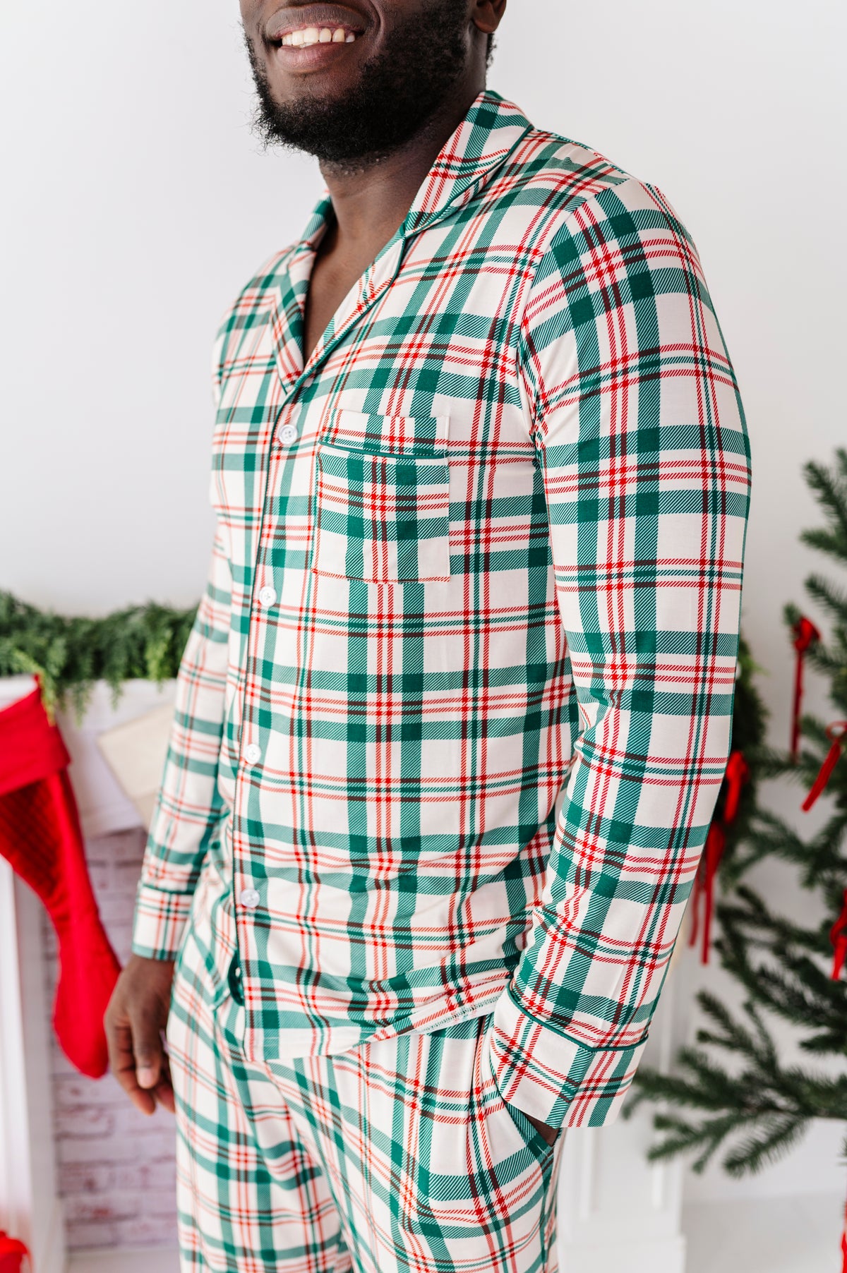 A side view of a man wearing a bamboo lounge set in a classic Christmas plaid print, standing in front of a fireplace and a Christmas tree