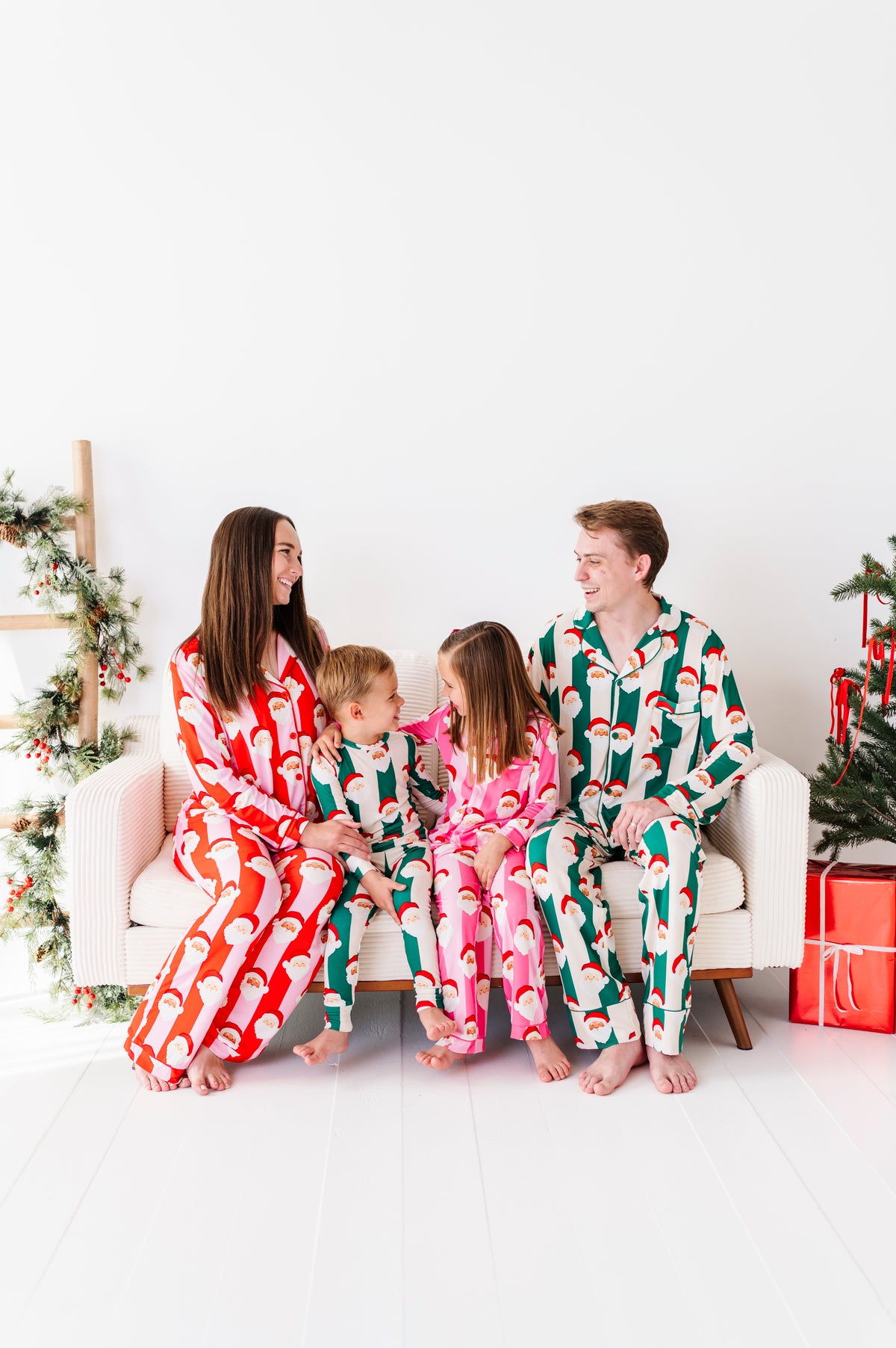 A family of four wearing matching Christmas-themed bamboo pajamas sits together on a white sofa in a bright, modern room decorated with a Christmas tree and wrapped gifts.