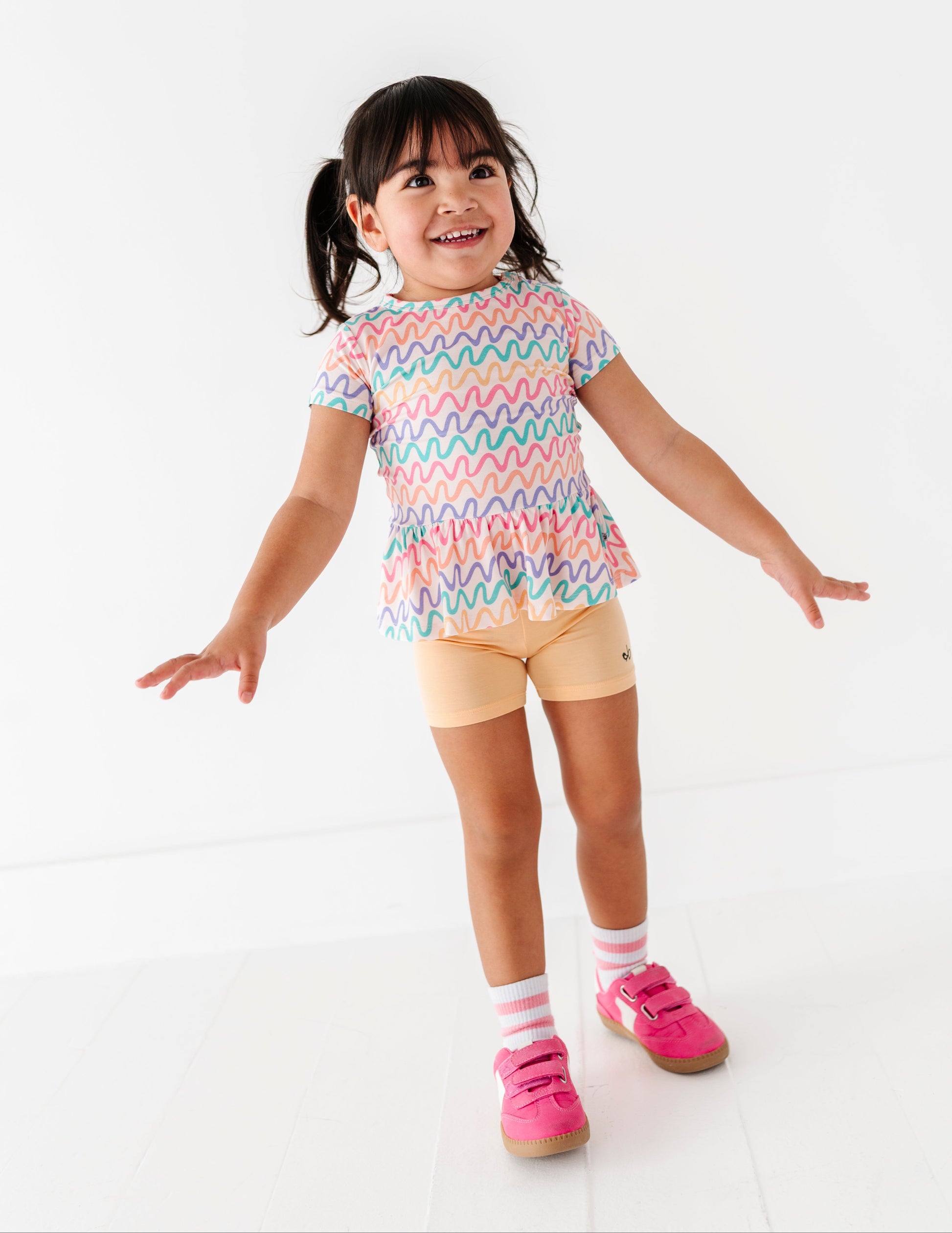 Child wearing a colorful wavy patterned bamboo/cotton peplum top and yellow bamboo biker shorts on a white background