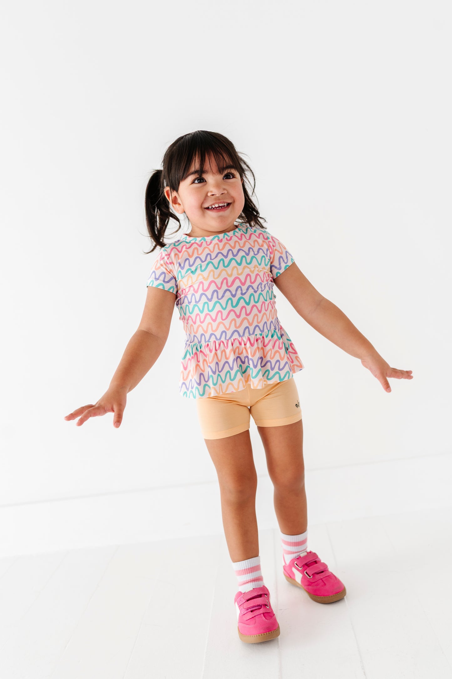 Child wearing a colorful wavy patterned bamboo/cotton shirt and butter yellow biker shorts on a white background