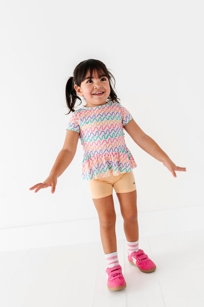 Child wearing a colorful wavy patterned bamboo/cotton shirt and butter yellow biker shorts on a white background