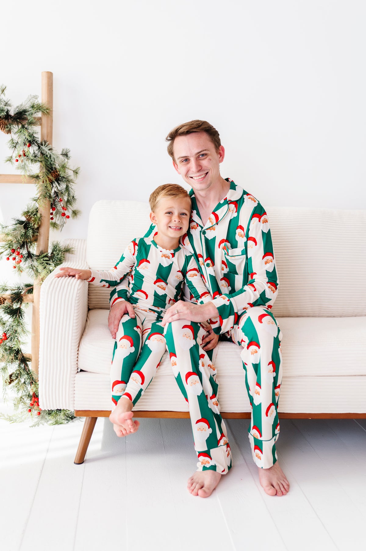 A man and a young boy wearing matching Santa Claus patterned bamboo pajamas sitting together on a white sofa in a bright, modern room.