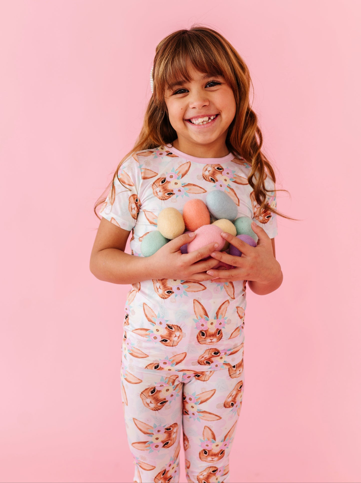 Young girl wearing a bunny-themed bamboo pajama set holding colorful eggs against a pink background