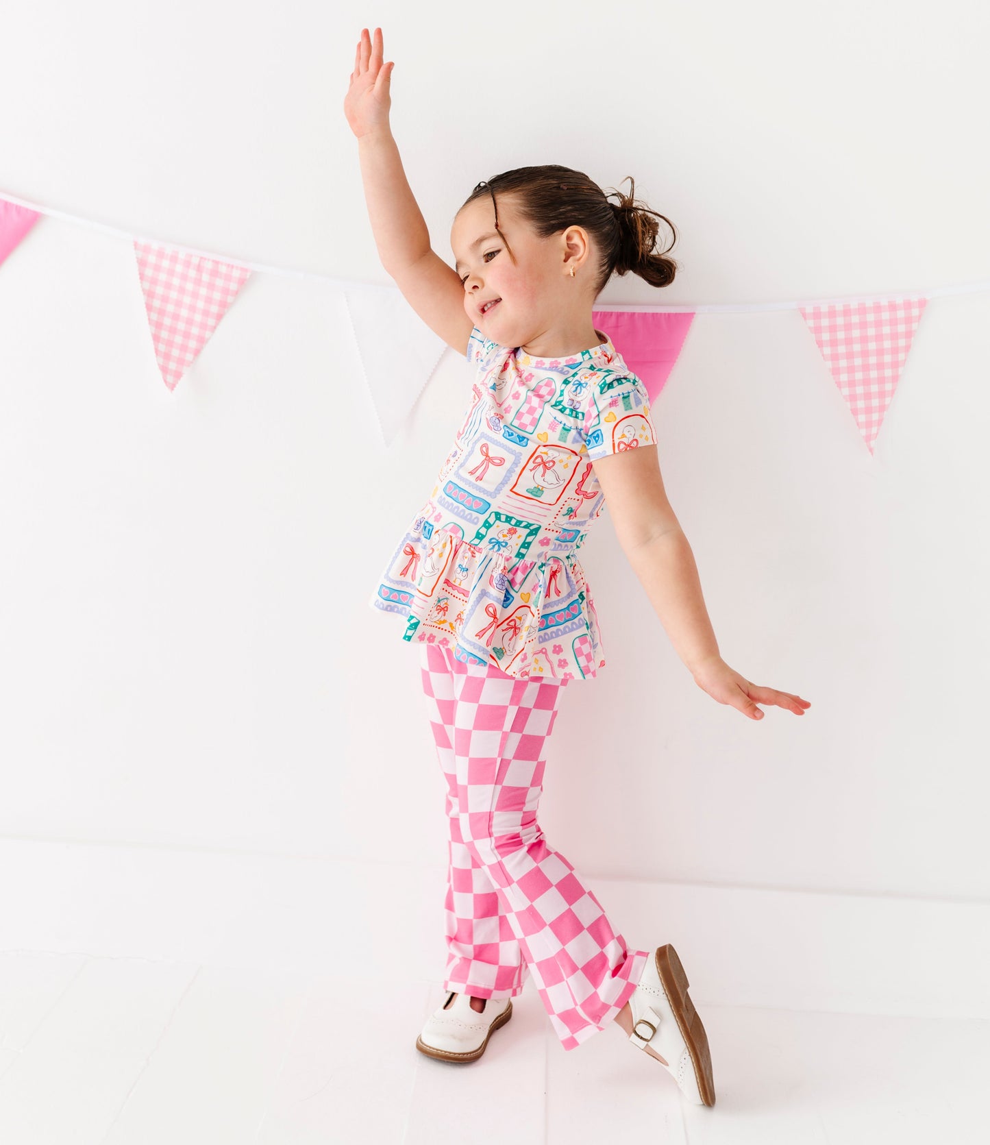 Little girl wearing a Gilly Goose Bamboo/Cotton Peplum Top with pink checkered pants against a white background