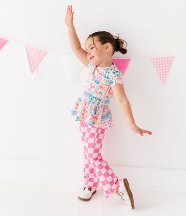 Little girl wearing a Gilly Goose Bamboo/Cotton Peplum Top with pink checkered pants against a white background