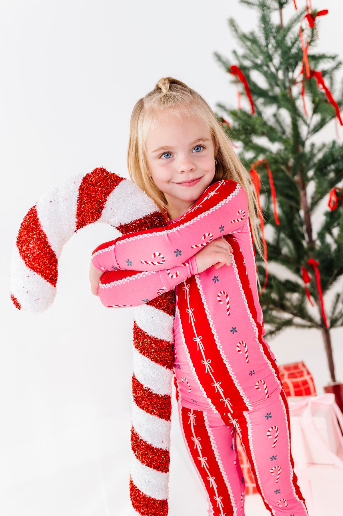 A young girl with blonde hair in a ponytail smiles while hugging a giant red and white striped candy cane plush toy. She is wearing a pink and red holiday bamboo two-piece set with candy cane patterns, standing in front of a decorated Christmas tree on a white background.