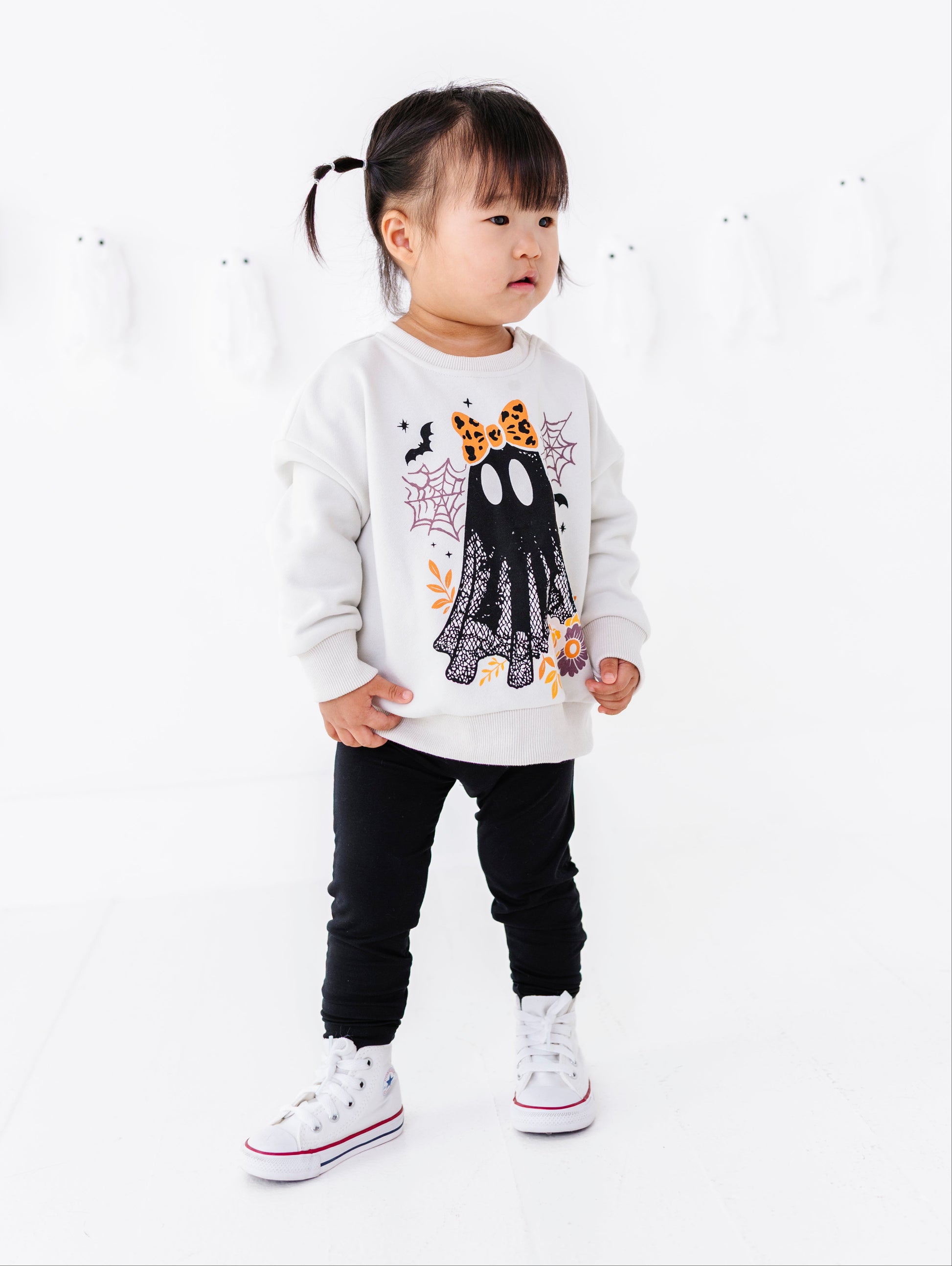 A toddler girl wearing a white long-sleeve sweatshirt with a black ghost graphic, black pants, and white sneakers, standing against a clean white studio background.