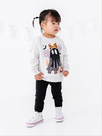 A toddler girl wearing a white long-sleeve sweatshirt with a black ghost graphic, black pants, and white sneakers, standing against a clean white studio background.
