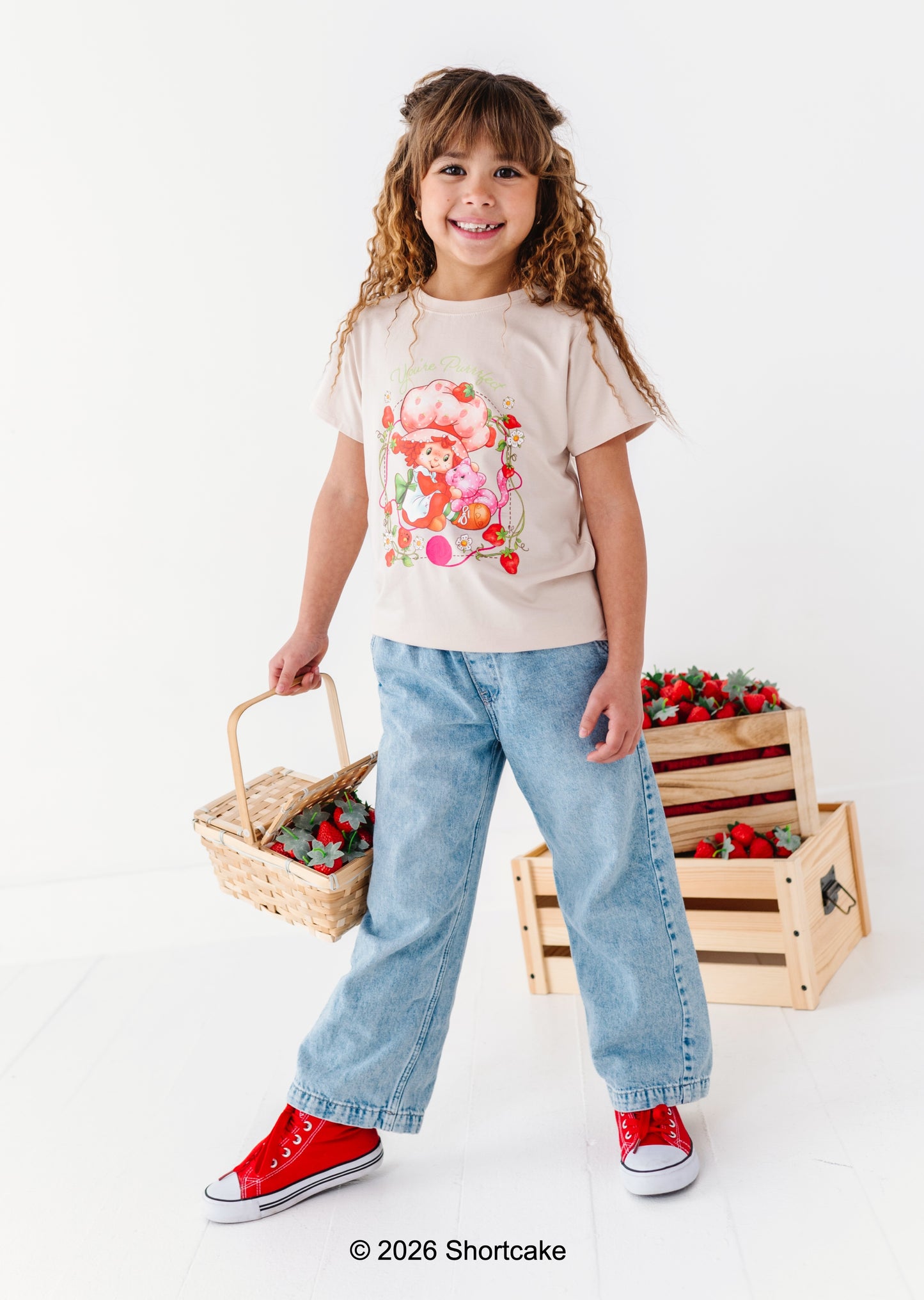 Young girl holding a basket with strawberries, wearing a Strawberry Shortcake™ t-shirt and jeans, on a white background.