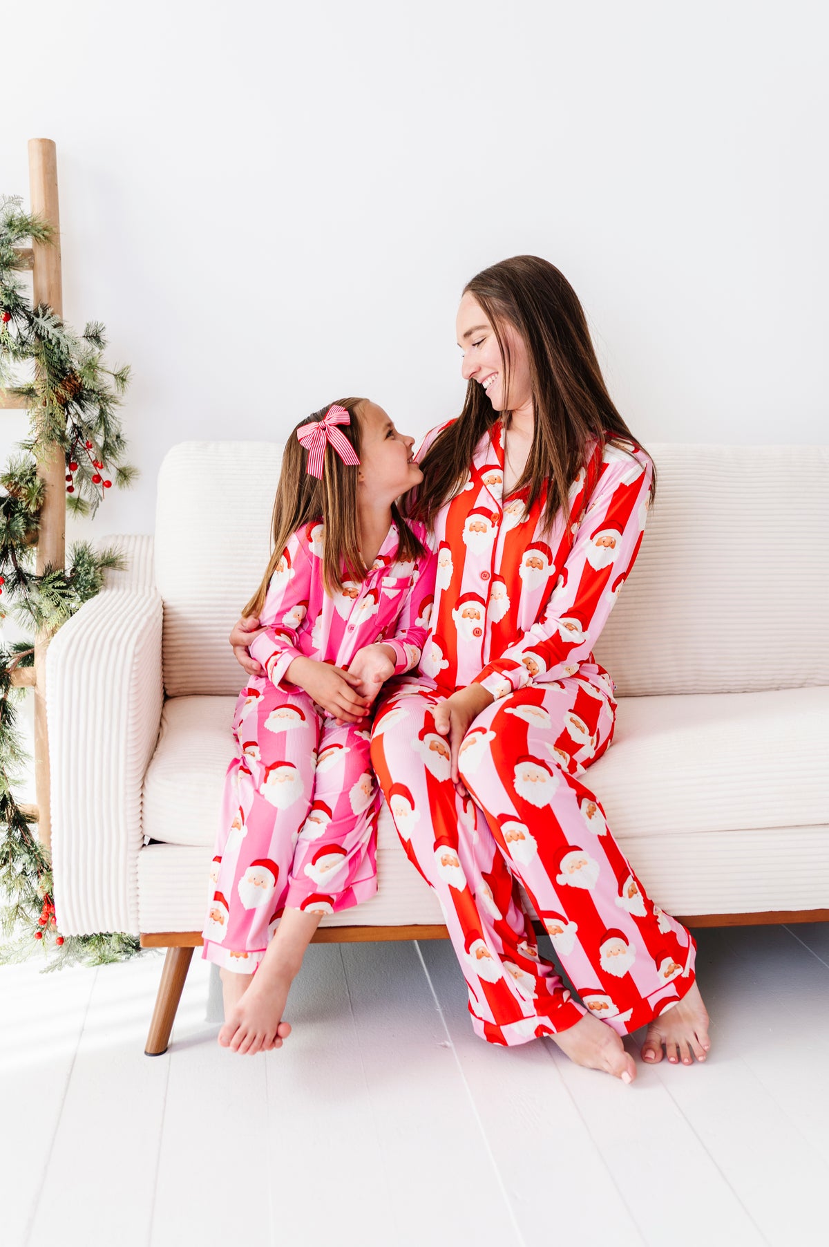 A woman and a young girl wearing matching Santa-themed bamboo pajamas sit together on a white sofa. The woman's pajamas are red with a white Santa pattern, and the girl's are pink with a white Santa pattern.