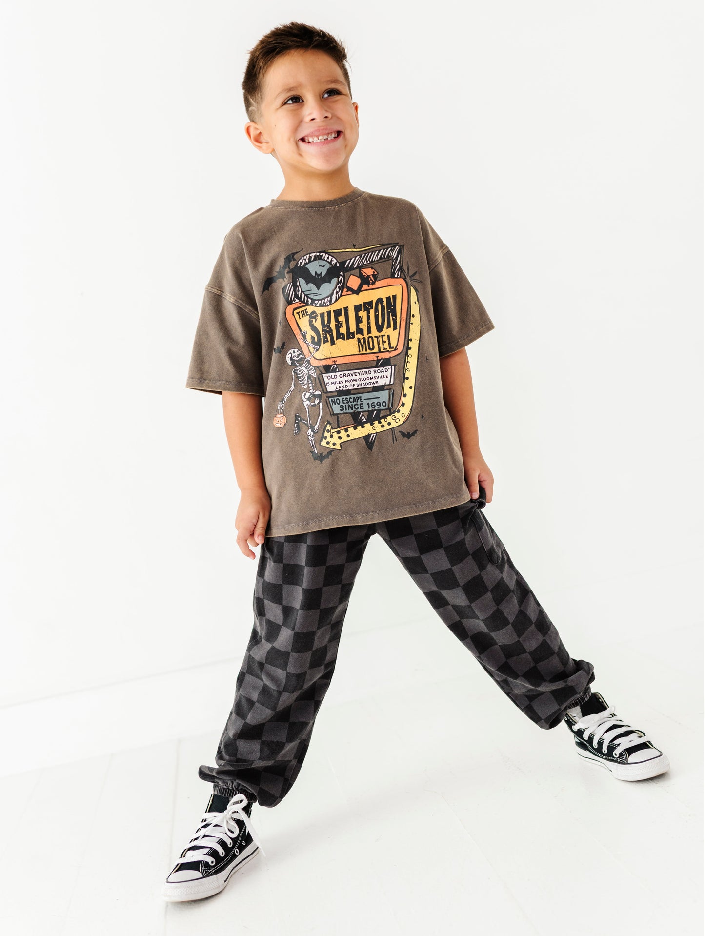 A young boy modeling a brown graphic t-shirt with a 'SKELETON MOTOR' design, paired with black and gray checkered pants and black-and-white sneakers, against a clean white studio background.