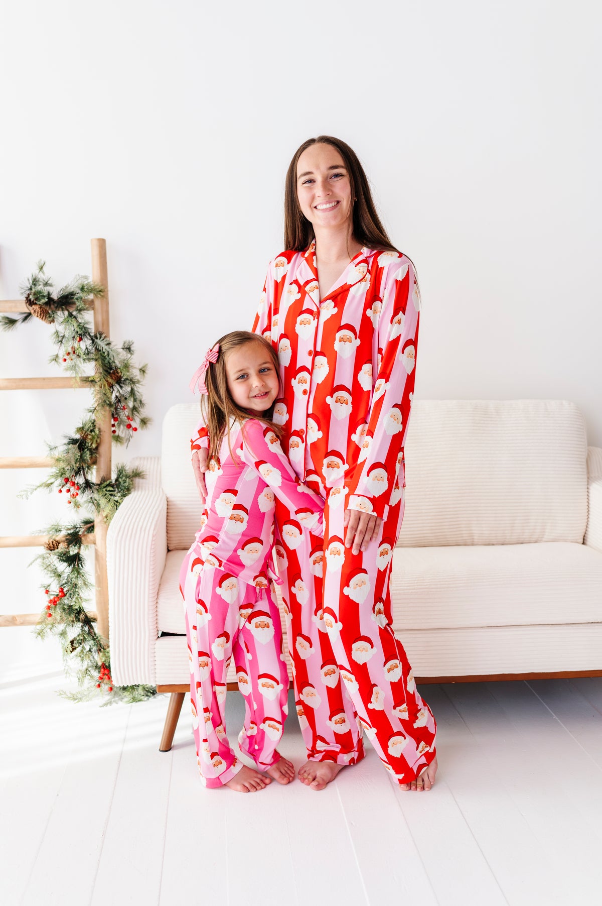 A woman and a young girl wearing matching Santa Claus patterned bamboo pajamas, standing together in a bright, modern living room with a white sofa and holiday greenery.